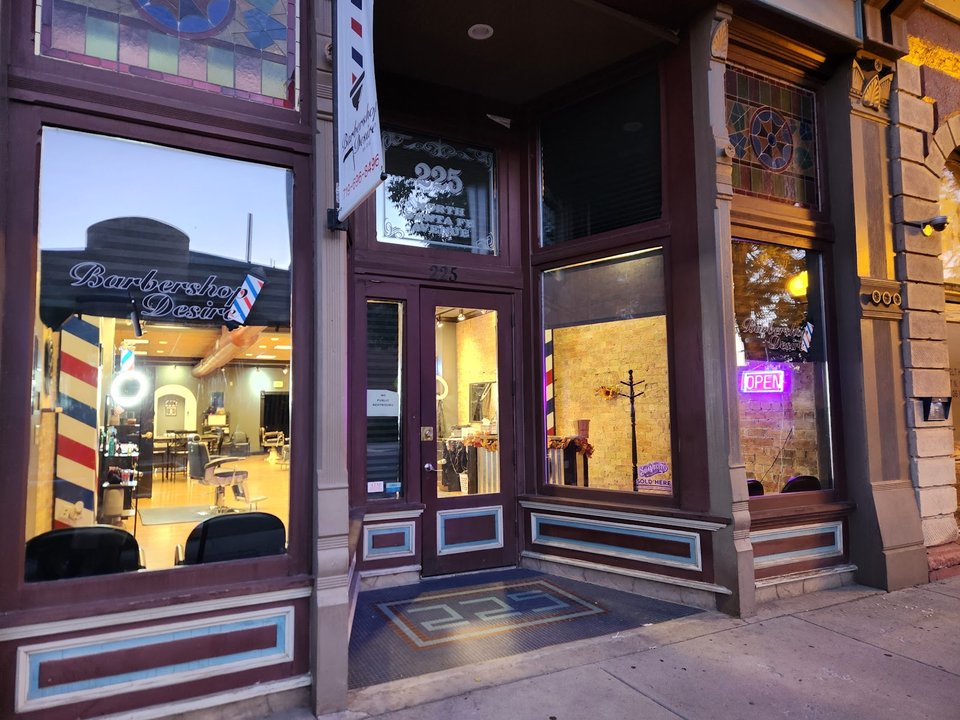 Street view of the Barbershop Desire storefront, stained glass, a barber pole, and chairs visible through the window.
